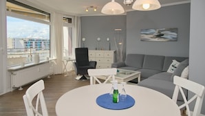 Uma sala de estar moderna com um sofá cinzento, cadeiras brancas, uma mesa de centro de vidro e uma mesa de jantar com um jogo americano azul. Há uma janela com vista para edifícios e um radiador abaixo.