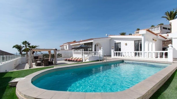 A swimming pool surrounded by a white building with a curved edge, a covered seating area, and palm trees. - Villa Melua (Chayofa)