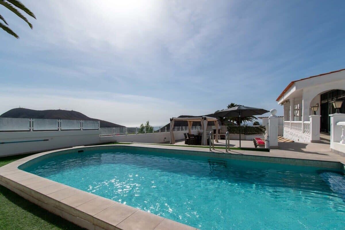 Ein Swimmingpool mit einer weißen Poolterrasse, einem überdachten Sitzbereich und einem Gebäude mit weißer Fassade.