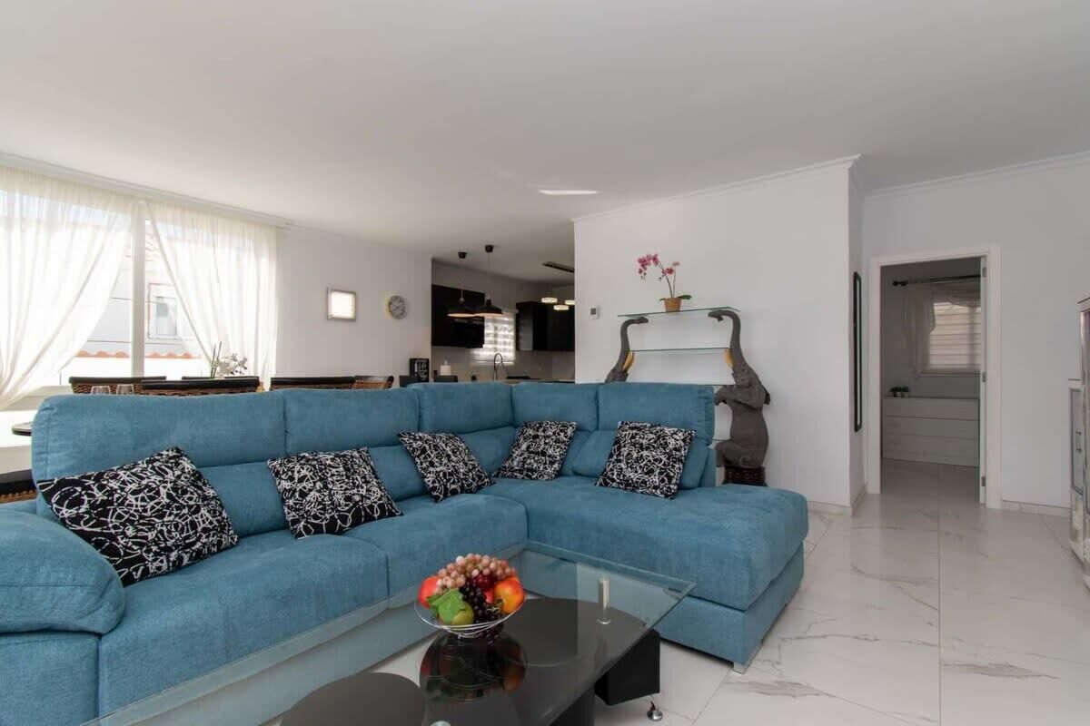 Ein modernes Wohnzimmer mit einem blauen Ecksofa, einem Glastisch mit Obst und einer minimalistischen Einrichtung.
