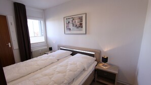 A hotel room with a double bed, a nightstand with a lamp, a framed picture on the wall, and a window with curtains. - Grömitz-center II App. 41 Inkl. Wlan (Grömitz)