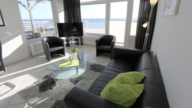 A modern living room with a black leather sofa, a glass coffee table, and a view of the sea through large windows. - Grömitz-center II App. 41 Inkl. Wlan (Grömitz)