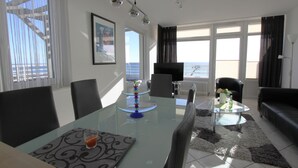 A modern living room with a glass dining table, black leather sofa, and a view of the sea through large windows. - Grömitz-center II App. 41 Inkl. Wlan (Grömitz)