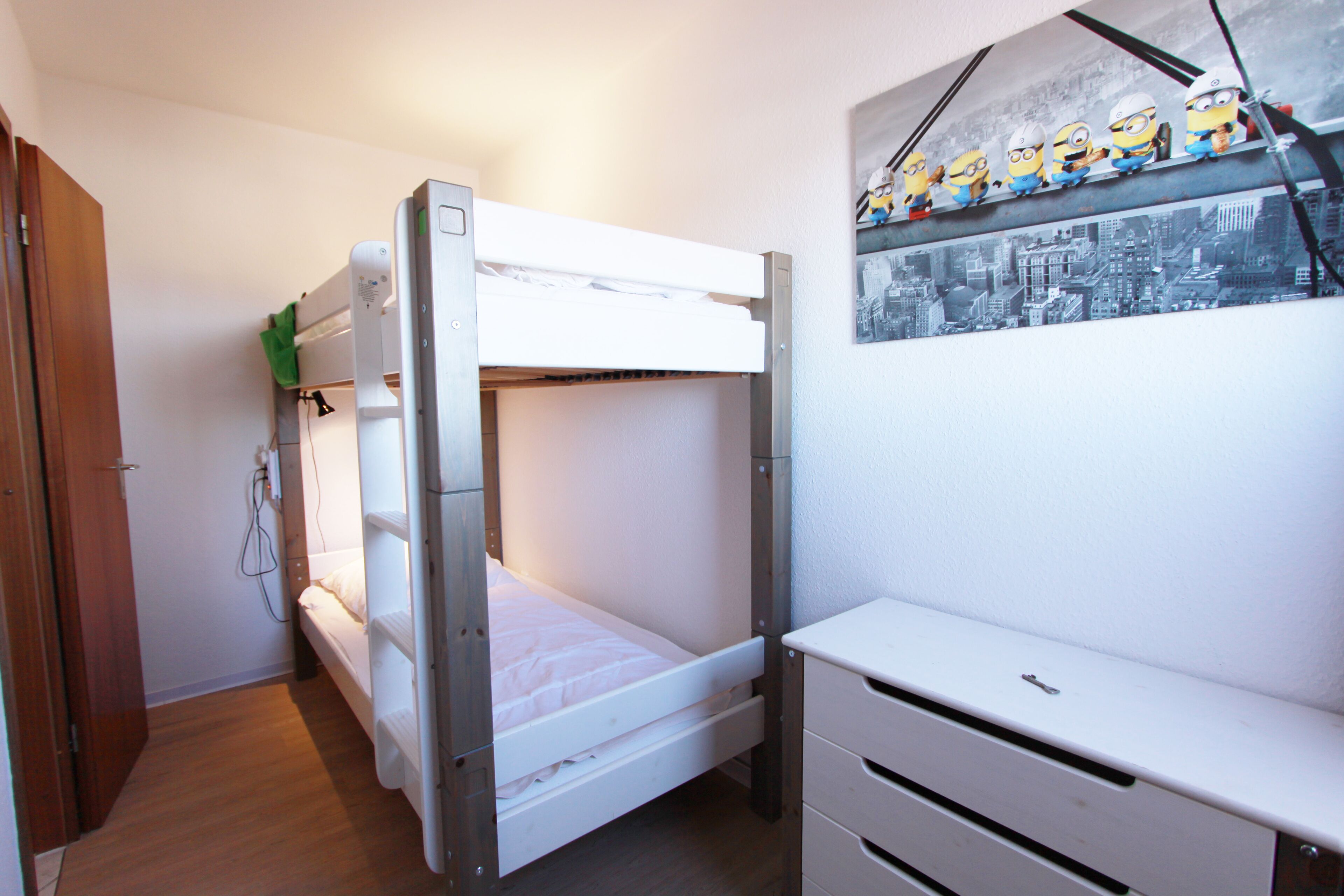 Un lit superposé avec un matelas blanc et un petit bureau doté de tiroirs. Il y a une étagère fixée au mur, sur laquelle sont posées des figurines, au-dessus du lit.