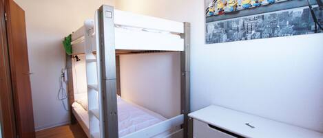 Un lit superposé avec un matelas blanc et un petit bureau doté de tiroirs. Il y a une étagère fixée au mur, sur laquelle sont posées des figurines, au-dessus du lit.