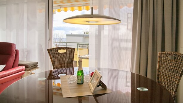 A hotel room with a round glass table, a red armchair, two wicker chairs, a bottle of champagne, and a framed picture. - Miramar App. 705 Inkl. Wlan (Grömitz)