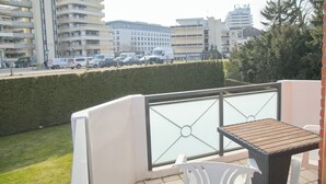 Un balcon doté d’une balustrade en verre, une petite table et des chaises. En arrière-plan, on aperçoit des immeubles à plusieurs étages et des voitures garées.