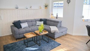 Un salon moderne avec un canapé d’angle gris, deux petites tables d’appoint en bois et un tapis bleu.