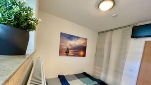 Une petite pièce comprenant un lit, un radiateur, une plante en pot, un tableau représentant un coucher de soleil et une fenêtre avec des stores.