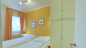 Un dormitorio con cama, armario, ventana con cortinas y dos cuadros en la pared.