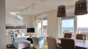A modern living room with a sofa, chairs, a dining table, and a TV. There is a large window with a view of the sea.