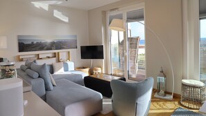 A modern living room with a sofa, a television, and a view of the sea through large windows.