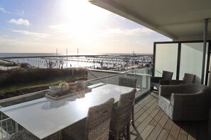 A balcony with a glass table and wicker chairs overlooking a coastal area with a pier. - Seeblick Seeloft Inkl. Wlan (Grömitz)