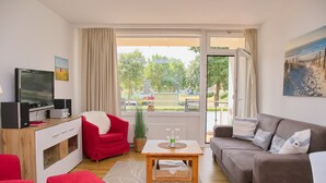 En moderne stue med et fjernsyn, røde og grå sofaer, et træbord og udsigt til en park og en gade.