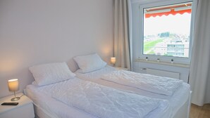 A double bed with white bedding, a bedside table with a lamp, and a window with a view of buildings. - Hanseat I Appartement. 79 (Grömitz)