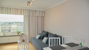 A modern living room with a grey sofa, a small white table, and a view of the city through large windows. - Hanseat I Appartement. 79 (Grömitz)