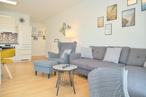 A modern living room with a grey sofa, a blue armchair, and a small round table. There is a kitchen area with white cabinets and a yellow chair. The walls are decorated with framed pictures and a clock. - Weiße Villa Whg. 5 Inkl. Wlan (Grömitz)