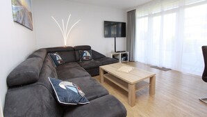 Un salon moderne avec un canapé d’angle, une table basse, un téléviseur à écran plat et une grande fenêtre agrémentée de rideaux.