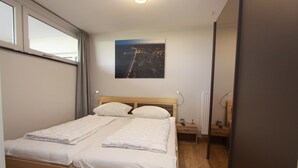 Un lit double bien fait, avec du linge de lit blanc, une tête de lit et une table de chevet sur laquelle se trouvent une lampe et une petite table.