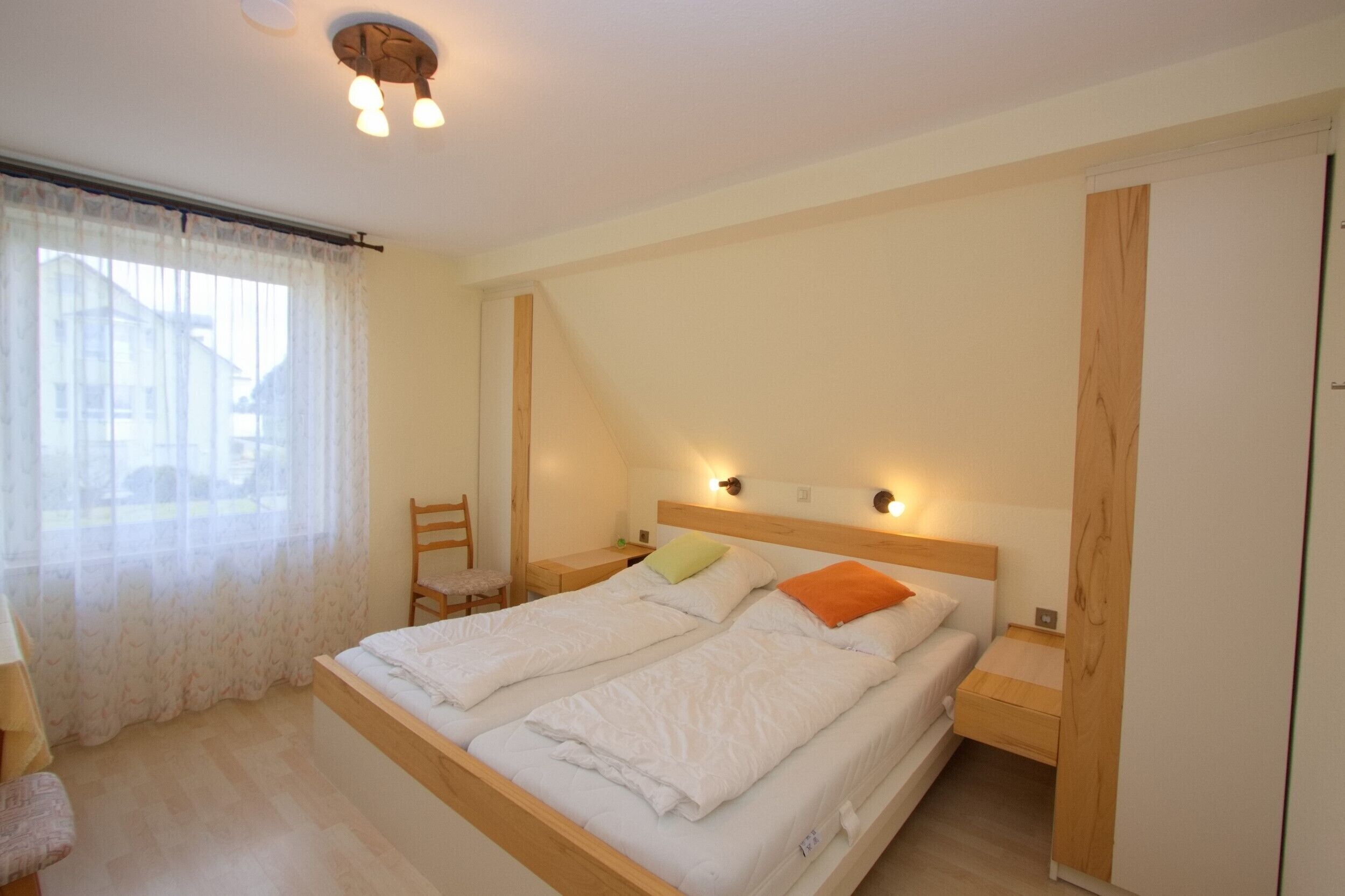 Un dormitorio con dos camas individuales, cabecero de madera, mesitas de noche, una silla y una ventana con cortinas de gasa.