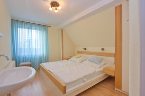 A bedroom with a bed, bedside tables, a nightstand, a window with curtains, and a bathroom with a sink and mirror.