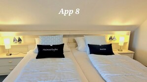 Habitació d'hotel amb dos llits, cadascun amb un coixí etiquetat "Heimathafen" i "Seerose". Hi ha làmpades de nit i una taula petita amb una làmpada a cada costat.