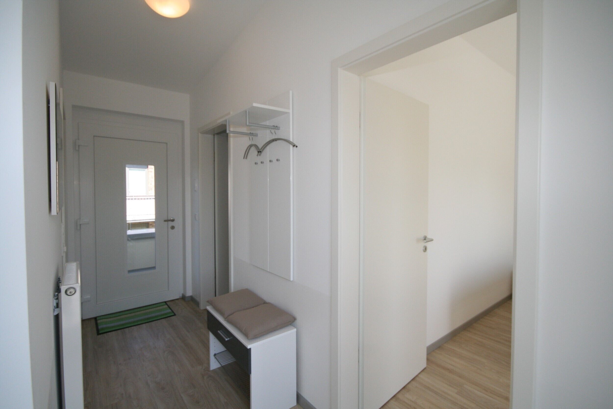 A hallway with a door leading to a room, a small bench, a coat rack, and a mirror on the wall.
