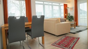 A modern living room with a sofa, two chairs, a dining table, and a large window with curtains. - Haus Belvedere App. 1 Inkl. Wlan (Grömitz)