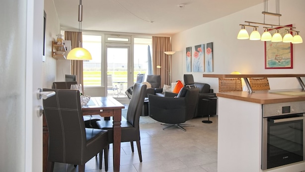 Et moderne køkken med indbygget ovn, et spiseområde med bord og stole, og en opholdsstue med sofaer og et fjernsyn.
