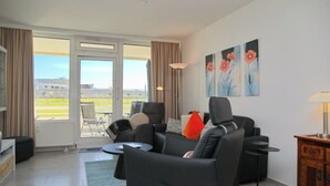 En moderne stue med sofa, lænestole, sofabord og fjernsyn. Der er to billeder på væggen, et vindue med gardiner og udsigt udad.