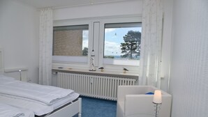 A hotel room with a bed, a small table with a lamp, a window with curtains, and a view of the outside. - Stettiner Str. 55 Inkl. Wlan (Grömitz)