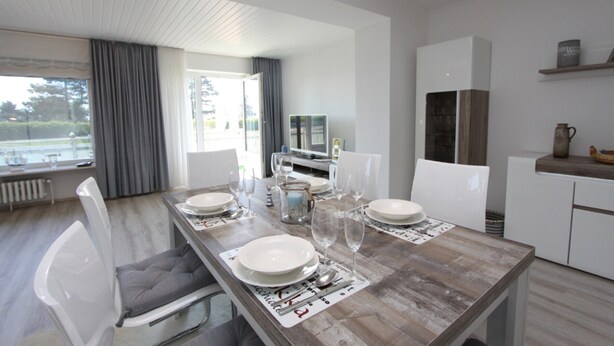 A modern dining area with a wooden table set for four, white chairs, and a view of the outdoors through large windows. - Stettiner Str. 55 Inkl. Wlan (Grömitz)