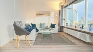 A modern living room with a white sofa, a glass coffee table, and a grey chair. There is a large window with a view of the city. - Stettiner Str. 55 Inkl. Wlan (Grömitz)