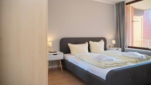 A hotel room with a bed, bedside tables, a window with curtains, and a view of a building outside. - Haus Belvedere App. 26 Wlan Inkl (Grömitz)