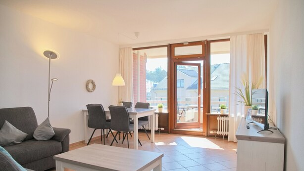 A living room with a sofa, coffee table, dining table with chairs, a lamp, and a view through a glass door to the outside. - Haus Belvedere App. 26 Wlan Inkl (Grömitz)