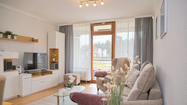 A modern living room with a sofa, a glass coffee table, a television, and a balcony with curtains. - Haus Belvedere App. 28 Inkl. Wlan (Grömitz)