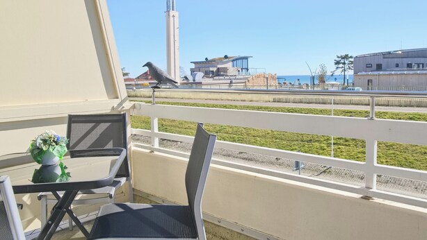 Un balcon avec une table et des chaises, une balustrade et une vue sur un immeuble de grande hauteur et une étendue d’eau.