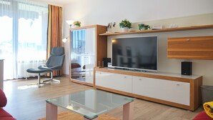 Un salon moderne avec une table basse en verre, un fauteuil gris, un grand téléviseur à écran plat et un meuble de rangement en bois agrémenté d’objets décoratifs.