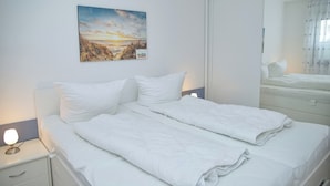 Un lit bien fait, avec du linge de lit blanc, une lampe de chevet, un tableau accroché au mur et un miroir qui reflète une partie de la pièce.