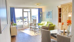 A modern living room with a green sofa, a dining table set for two, and a view through the glass doors to the outside. - Haus Belvedere App. 63 (Grömitz)
