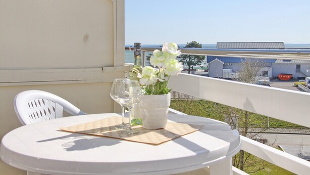 A balcony with a table, chairs, a vase of flowers, and a view of the sea and buildings. - Haus Belvedere App. 63 (Grömitz)
