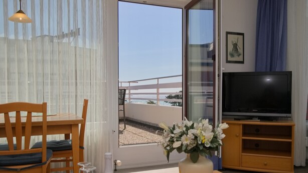 A room with a dining table, chairs, a TV, a vase with flowers, and a view of the sea through a sliding door. - Haus Belvedere App. 64 (Grömitz)