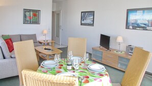 Un coin repas avec une table dressée pour quatre personnes, entouré de chaises en osier, d’un canapé et d’un téléviseur posé sur un meuble.