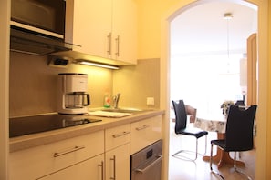 A kitchen with a coffee maker, sink, and cabinets. There is a dining area with a table and chairs visible in the background. - Haus Belvedere App. 73 Wlan Inkl (Grömitz)