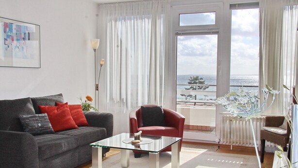 A living room with a grey sofa, red cushions, a glass coffee table, and a view of the sea through large windows. - Haus Belvedere App. 75 Wlan Inkl (Grömitz)