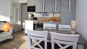 A modern kitchen with grey cabinets, a microwave, and a painting of a lighthouse. There is a dining table with white chairs and a candle on it. - Haus Belvedere App. 77 Wlan Inkl (Grömitz)