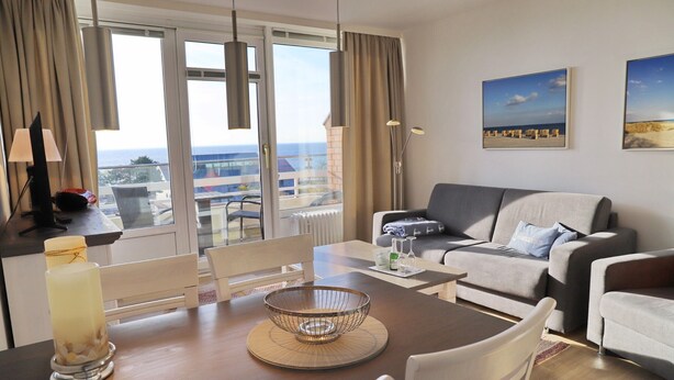 A modern living room with a dining area, featuring a large window with a view of the sea, a grey sofa, and a wooden dining table with a candle and a decorative bowl. - Haus Belvedere App. 77 Wlan Inkl (Grömitz)
