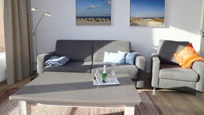 A modern living room with a grey sofa, a grey armchair, and a coffee table. There are two framed beach pictures on the wall. - Haus Belvedere App. 77 Wlan Inkl (Grömitz)