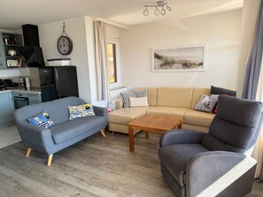 A living room with a grey sofa, a grey armchair, a wooden coffee table, a kitchen area with appliances, and a clock on the wall. - Haus Belvedere App. 19 Wlan Inkl (Grömitz)