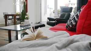 A modern living room with a glass coffee table, a red sofa, and a black armchair. There is a book on the sofa and a vase with flowers on the table.